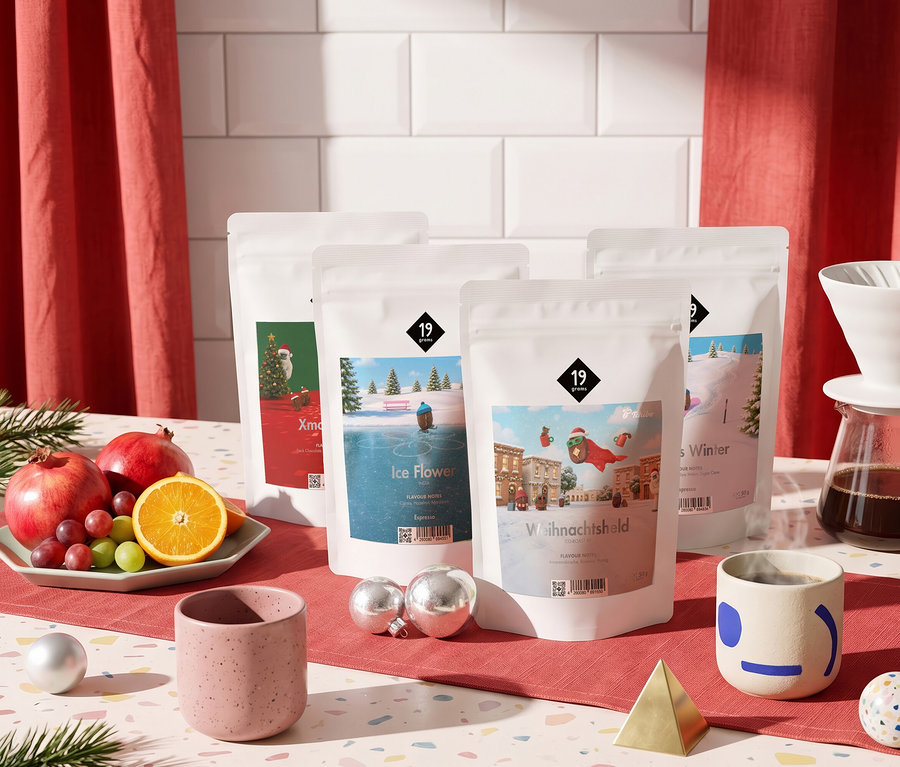 Fünf weiße Beutel mit winterlichen Motiven stehen auf einem roten Tischläufer, umgeben von Früchten, Kaffeegeschirr und Weihnachtsdekoration.