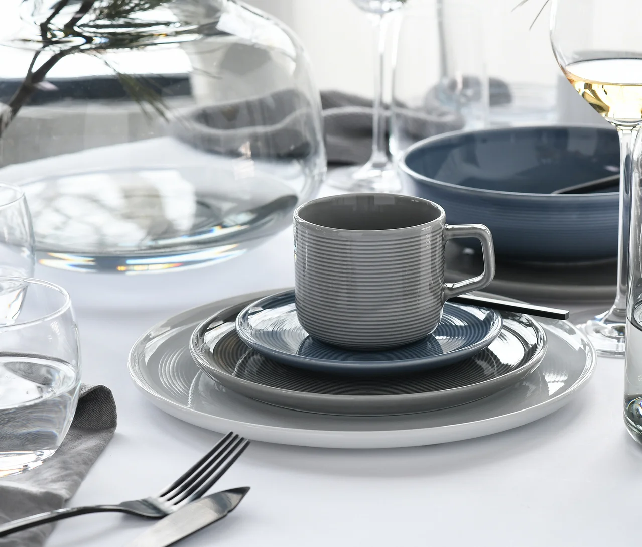 Ein Tisch ist mit einem creaTable Service »Session Urban« Geschirrset gedeckt, das einen Stapel weißer, grauer und blauer Teller, eine geriffelte graue Tasse und eine blaue Untertasse zeigt. Besteck, ein Wasserglas, eine Vase, eine blaue Schale und Weingläser sind ebenfalls sichtbar.