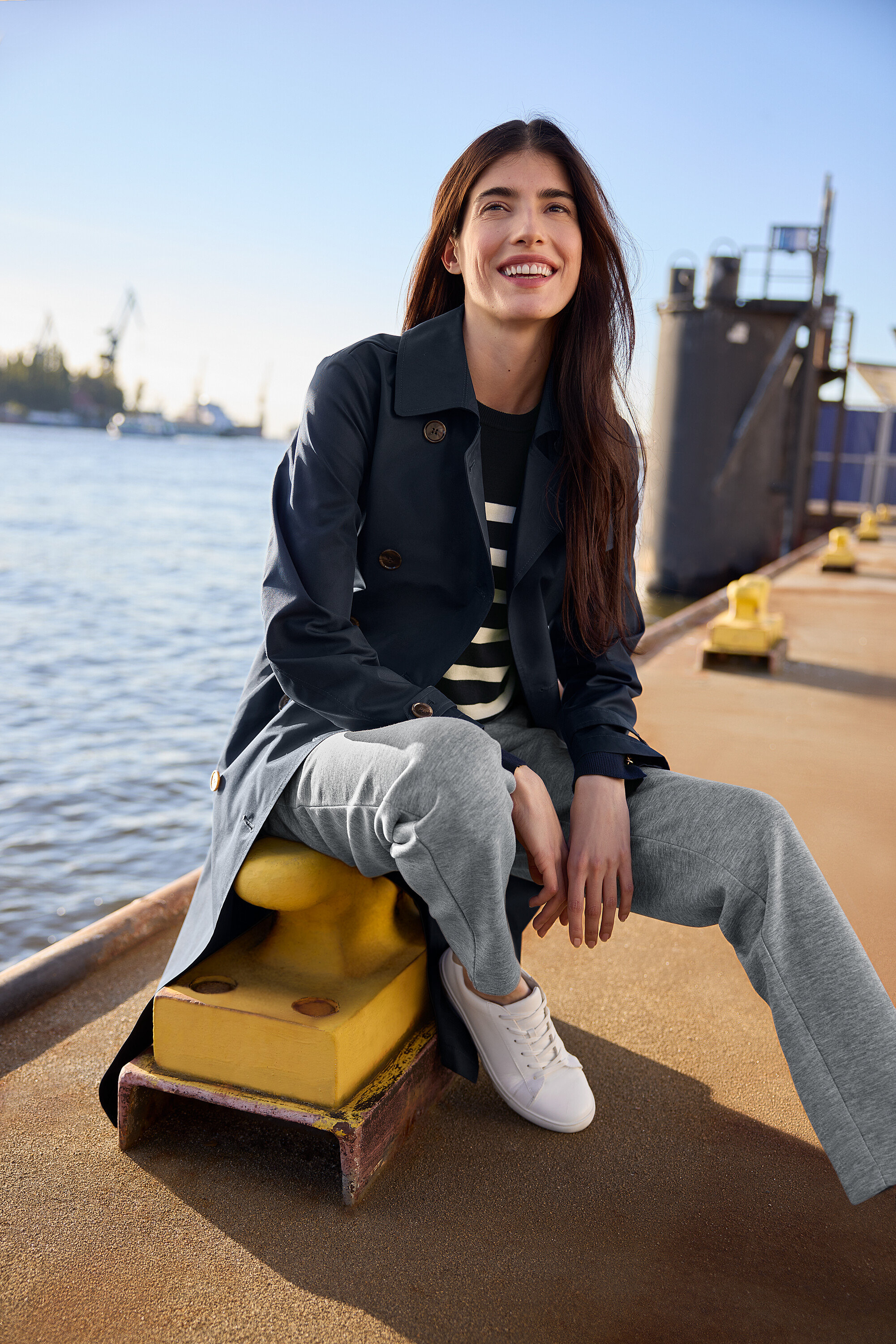 Lächelnde Frau sitzt auf einem gelben Poller und trägt einen Trenchcoat, einen gestreiften Feinstrickpullover, Joggpants und Leder-Sneaker.