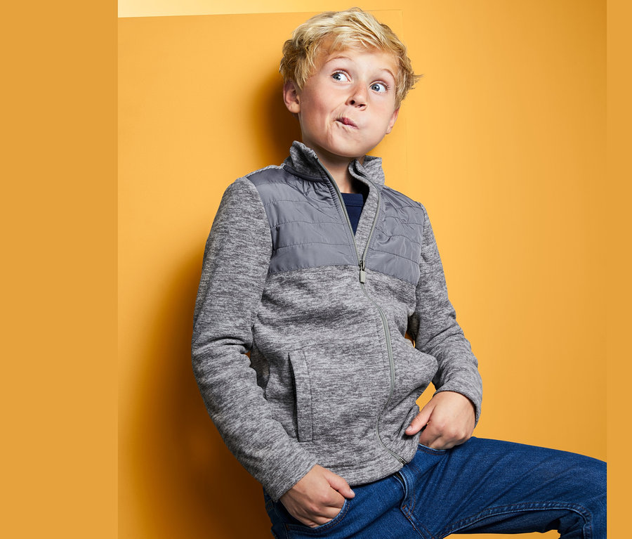 Junge posiert in Hybrid-Fleecejacke vor gelbem Hintergrund.