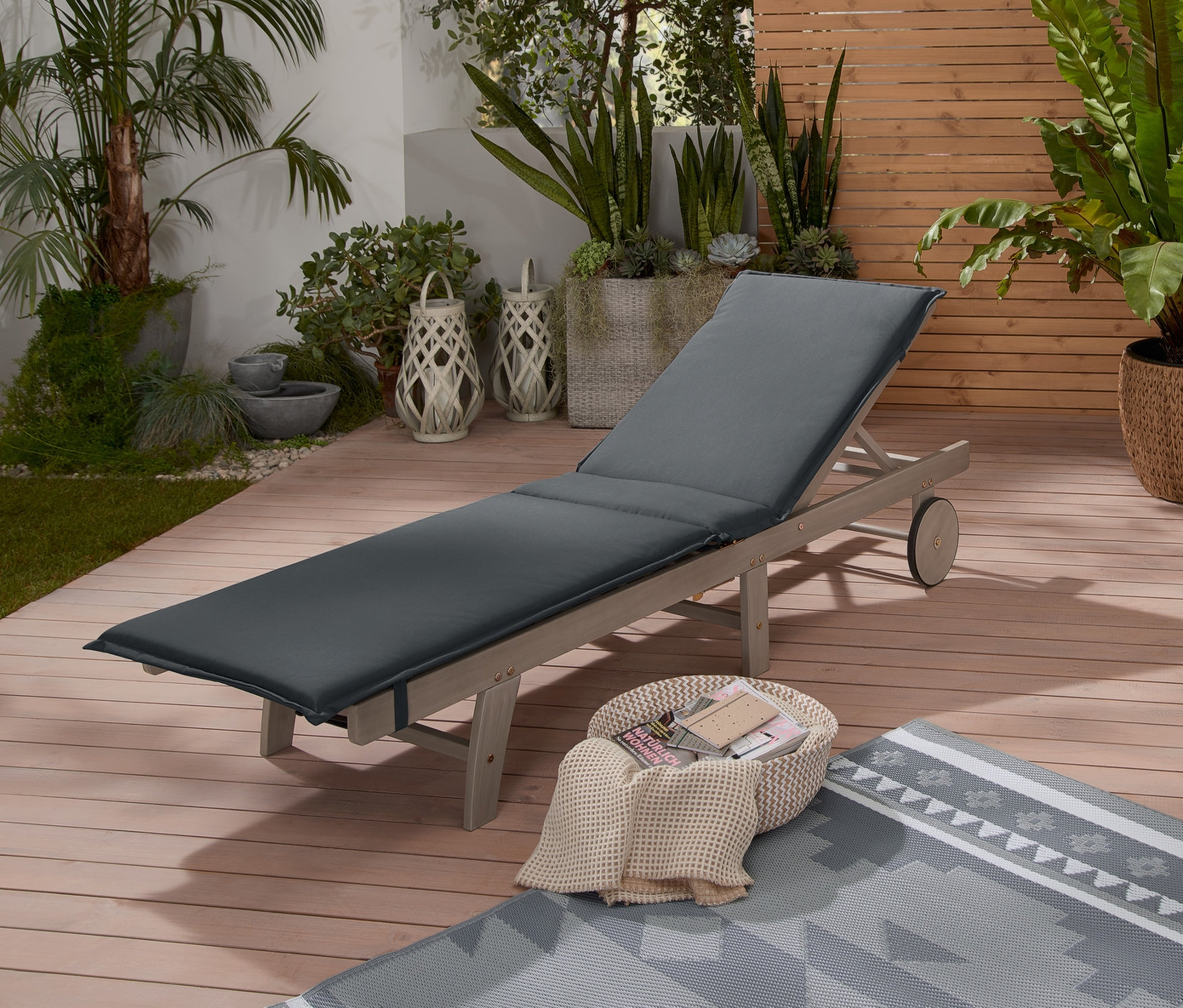 Eine klappbare Gartenliege in Vintage Grey steht auf einer Holzterrasse. Daneben liegen ein Outdoor-Teppich, ein geflochtener Korb und eine LED-Outdoor-Laterne mit Geflecht.