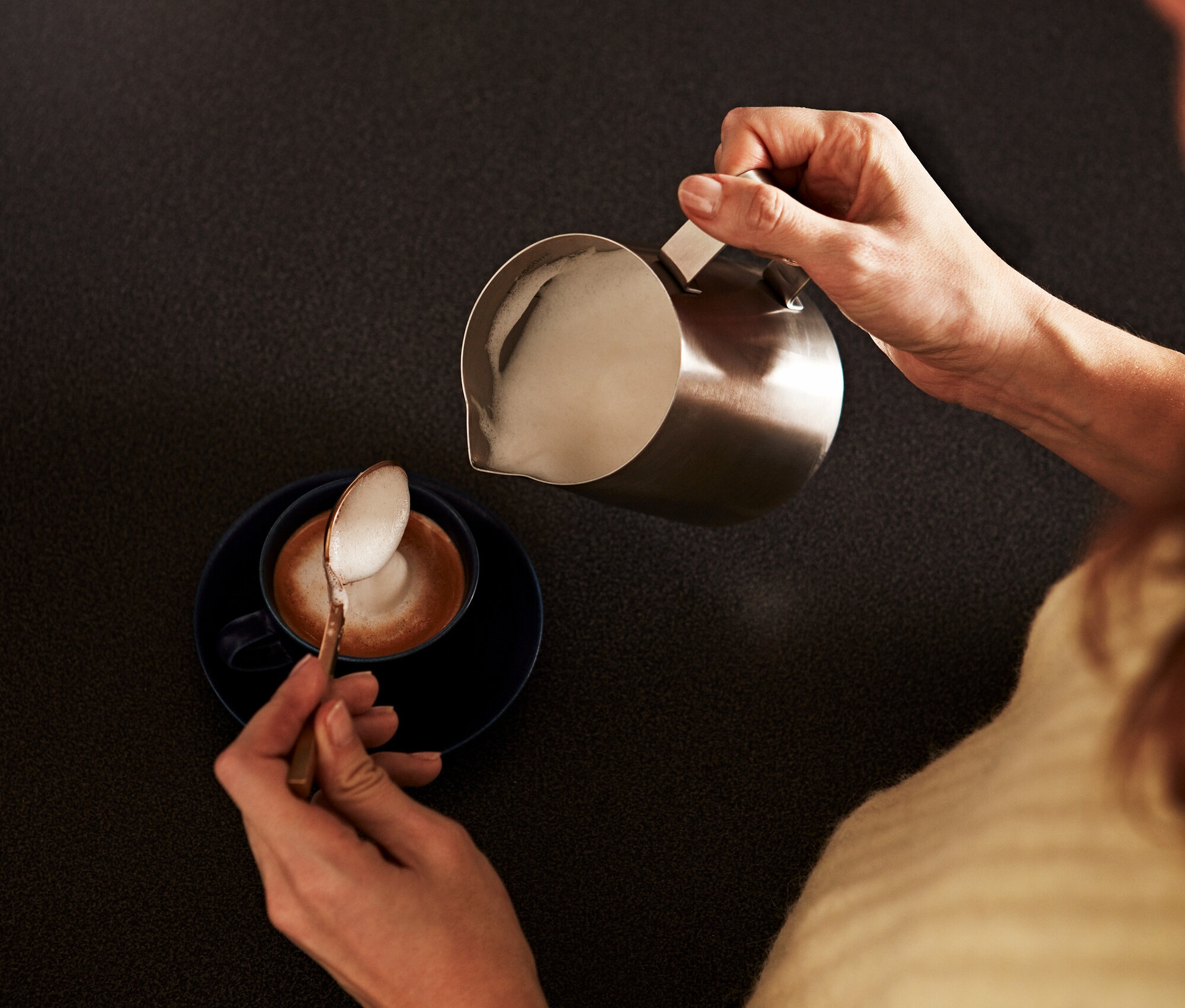 Frau gießt geschäumte Milch aus einem kleinen Edelstahl-Milchkännchen in eine Kaffeetasse und löffelt Schaum.