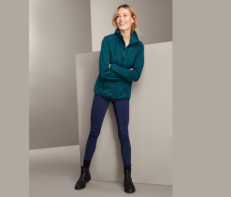 Frau posiert in Strickfleecejacke mit recyceltem Material, dunkelblauen Reithosen und schwarzen Stiefeln.