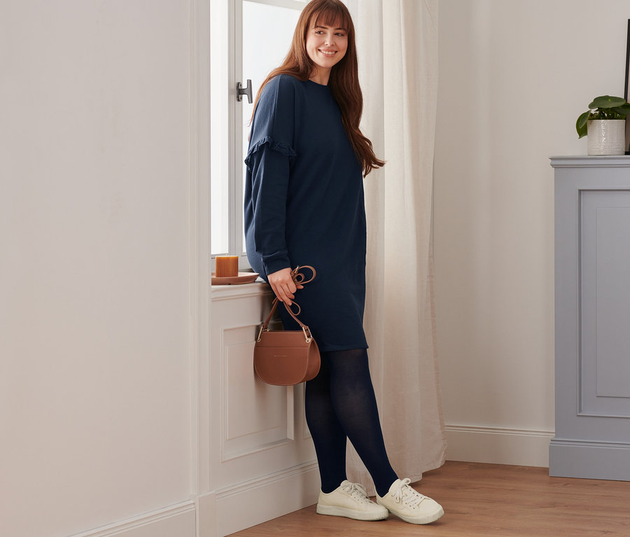 Eine lächelnde Frau lehnt am Fenster. Sie trägt ein Sweatkleid, Leder-Sneaker, eine blickdichte Strumpfhose (2er-Pack) und eine Umhängetasche.