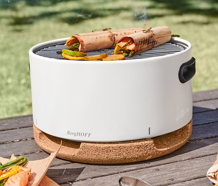Auf einem Tisch im Park steht ein BergHOFF® Tischgrill, auf dem Speisen in Wrap-Grillpapier aus Buchenholz gegrillt werden. Neben dem Grill liegen eine Edelstahl-Grillzange und Grillkohle-Anzünder.
