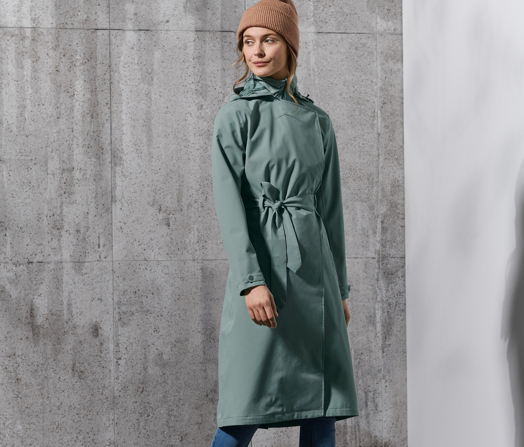 Frau posiert in einem grünen Regen-Trenchcoat, blauen Jeans, beigen Stiefeln und Mütze.