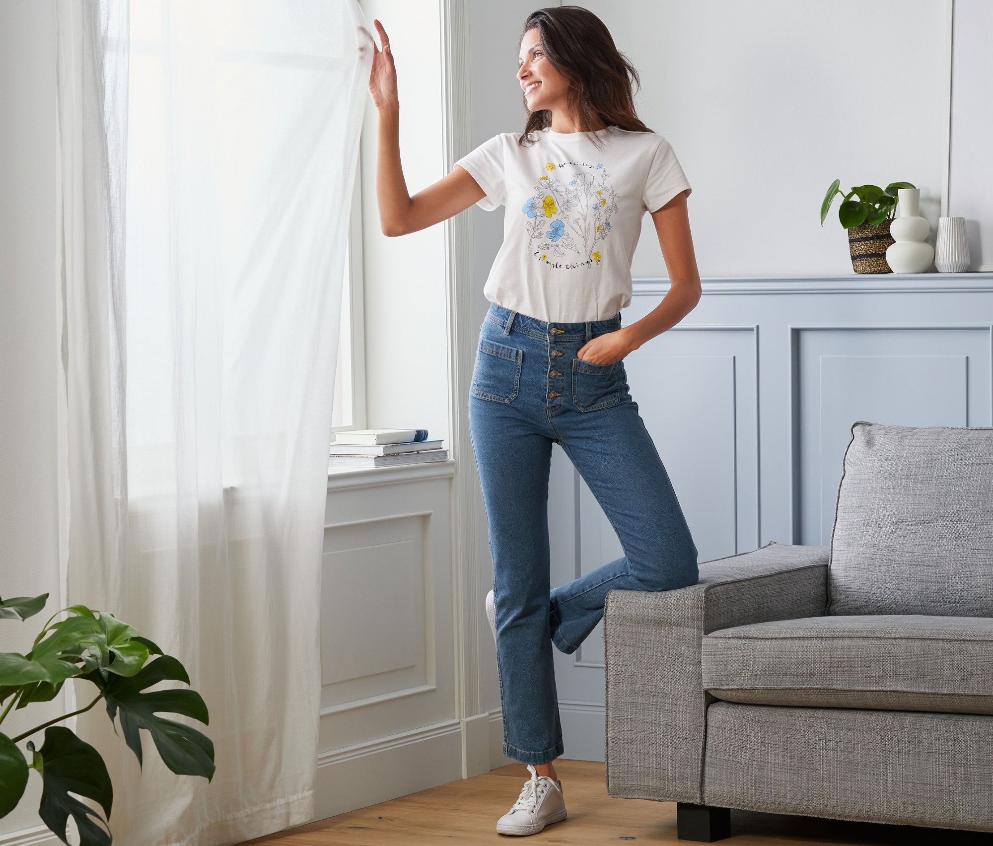 Eine Frau steht am Fenster und trägt Jeans mit Knopfleiste.