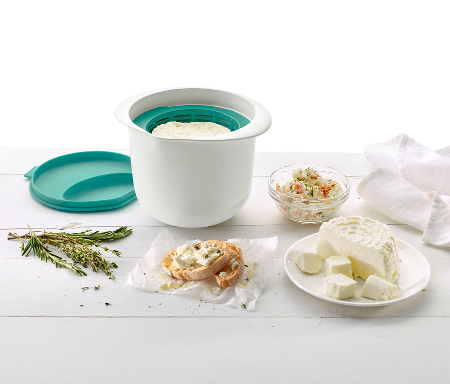 Das Bild zeigt ein Frischkäsebereiter-Set, ein Glas mit Aufstrich, Kräuter und einen Teller mit Frischkäse auf einem weißen Holztisch.