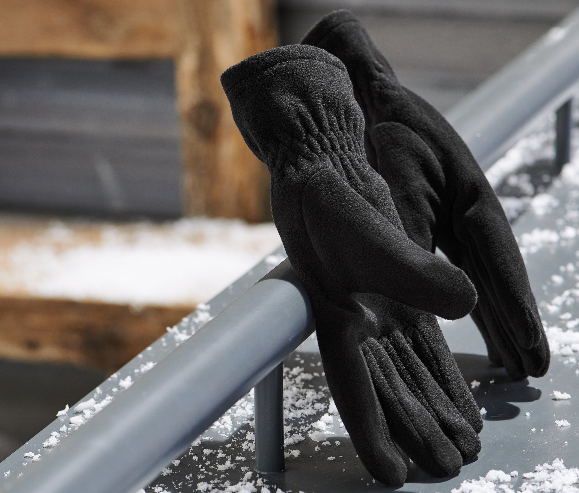 Schwarze Microfleece-Handschuhe liegen auf einer Metallstange. Im Hintergrund befindet sich eine Skibrille.