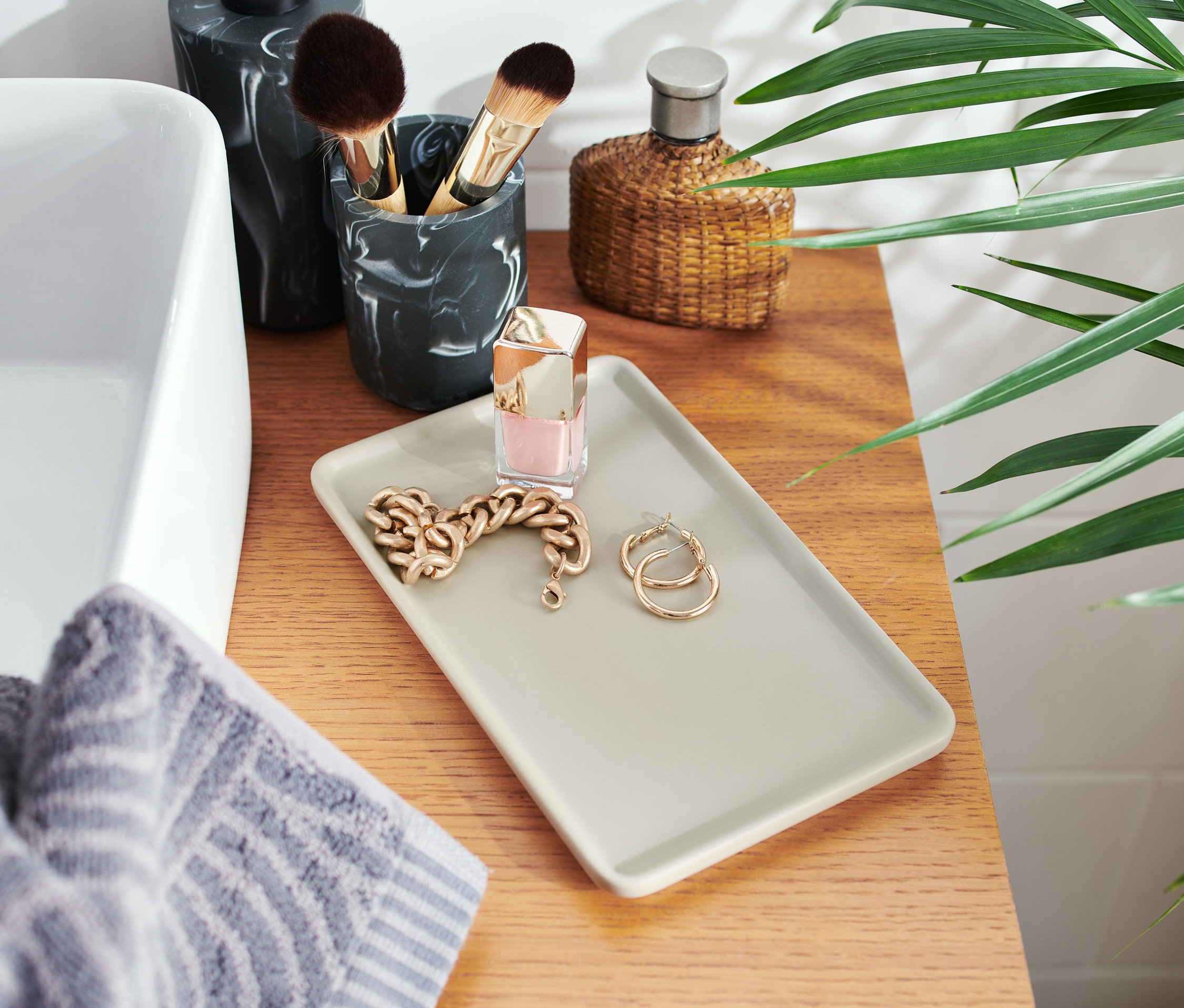 Keramik-Tablett mit Kette, Ohrringen und Nagellack auf einer Holzplatte neben einem Waschbecken.