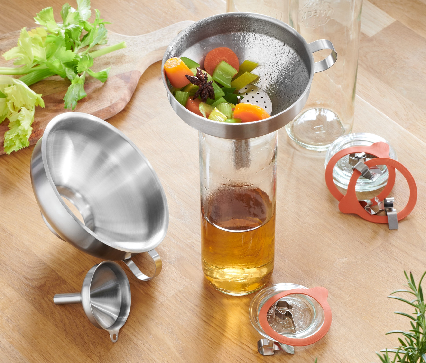 Edelstahltrichter-Set auf einem Holztisch mit Sellerie, Karotten, Gläsern und Gewürzen.