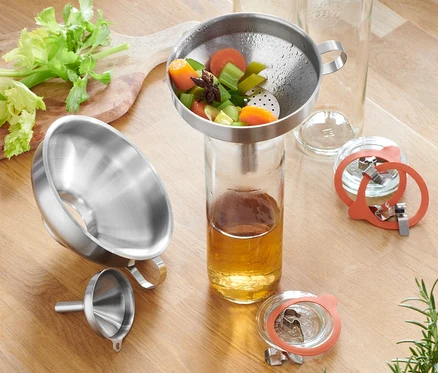 Edelstahltrichter-Set auf einem Holztisch mit Sellerie, Karotten, Gläsern und Gewürzen.