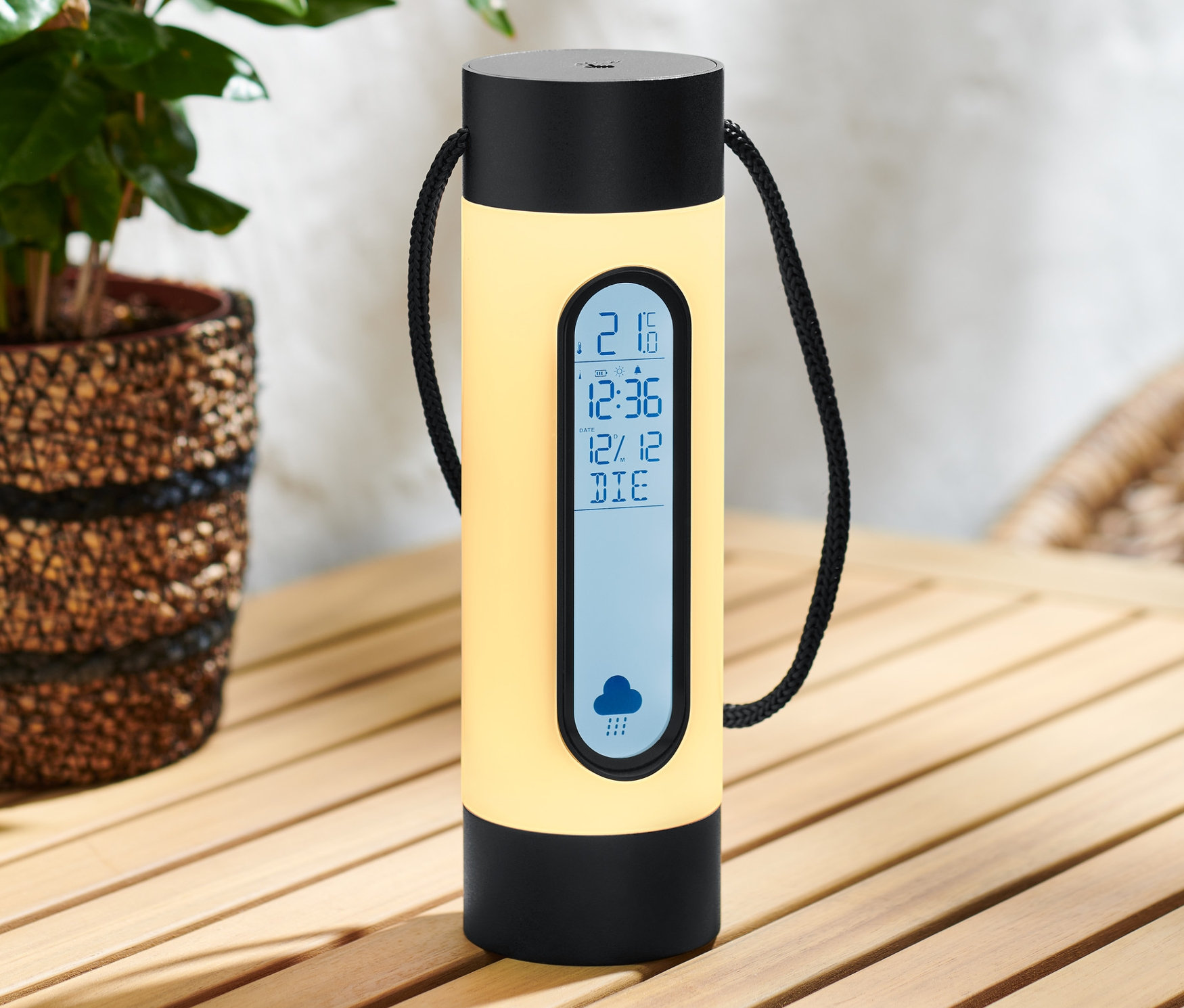 Eine gelbe Akku-Wetterstation mit Licht steht auf einem Holztisch neben einer Pflanze.