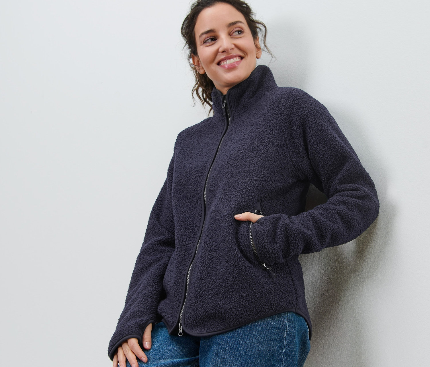 Lächelnde Frau lehnt an einer Wand und trägt eine marineblaue Teddyfleece-Jacke und Jeans.