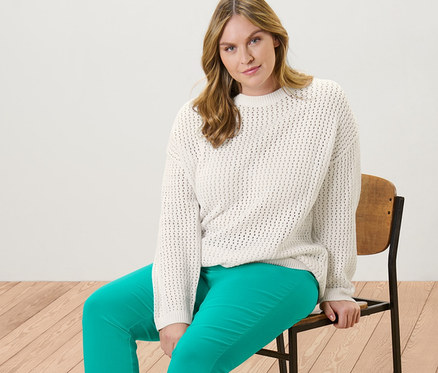 Frau sitzt auf einem Stuhl und trägt einen Pullover in Pointellestrick und eine 7/8-Stretchhose in Aqua.