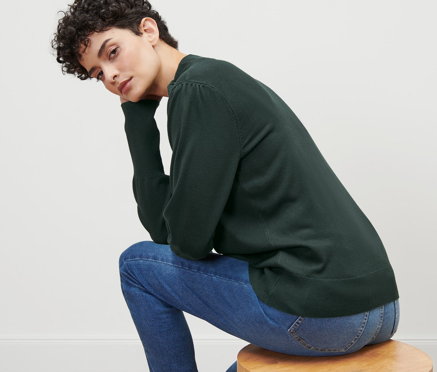 Frau sitzt auf einem Holzhocker und trägt einen grünen Feinstrickpullover mit Schleifendetail und Jeans.