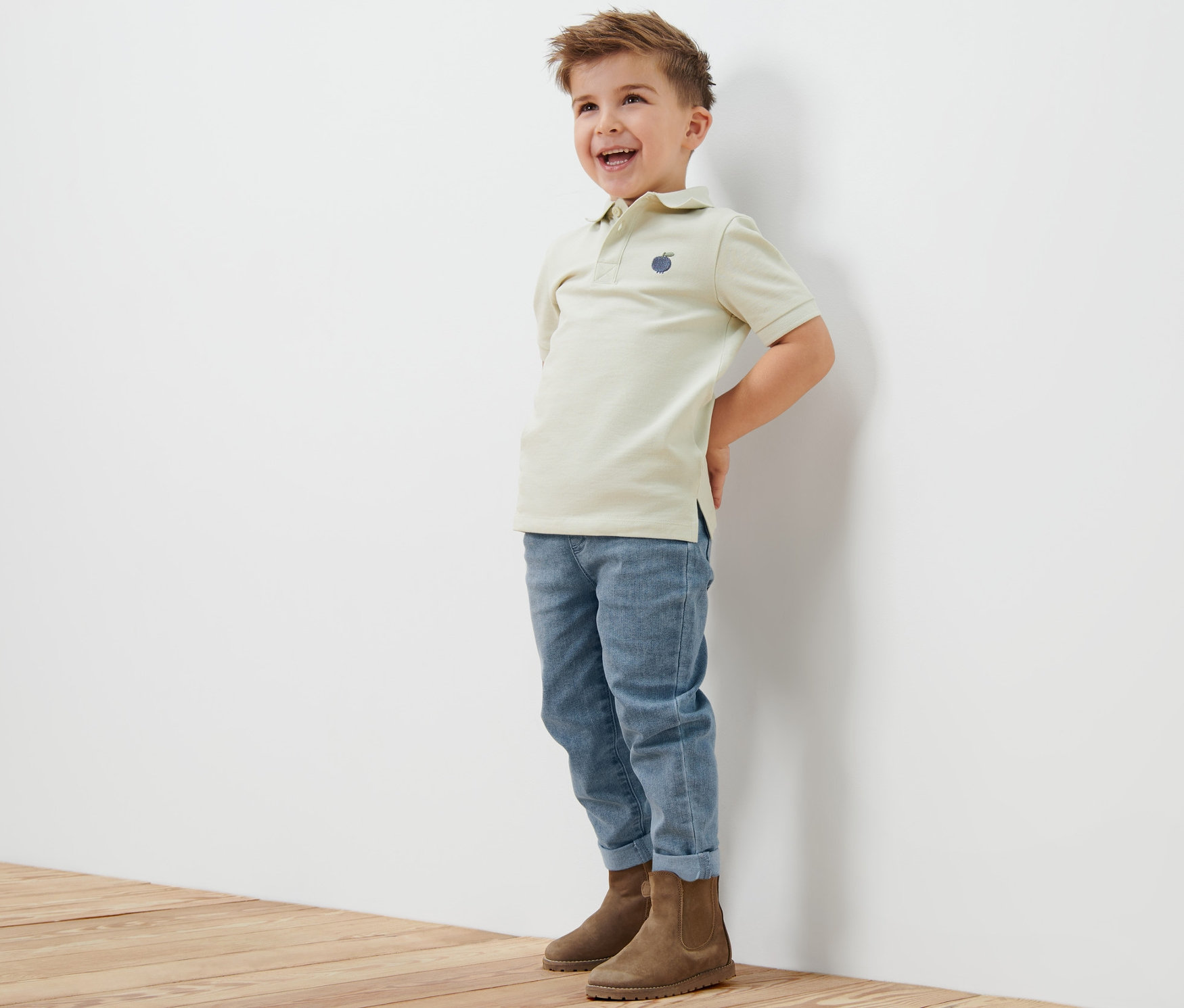 Ein lachender Junge steht an einer weißen Wand. Er trägt ein beiges Poloshirt, blaue Jeans und braune Stiefel. Die Jeans sind eine Kinder-Pull-on-Pants – Fit »Luca«.