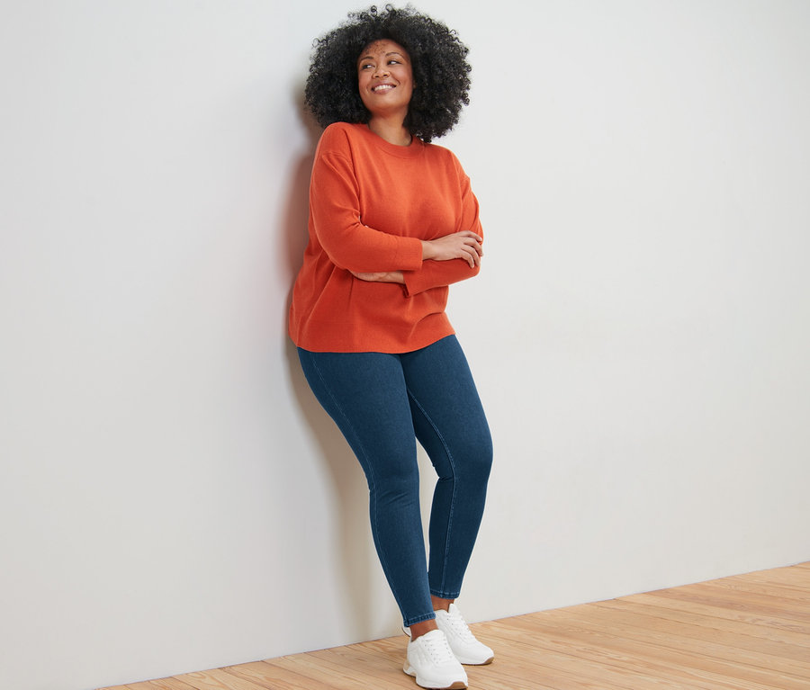 Eine Frau lehnt an einer Wand und trägt einen Feinstrick-Pullover mit Cashmere, Jeans und Turnschuhe.