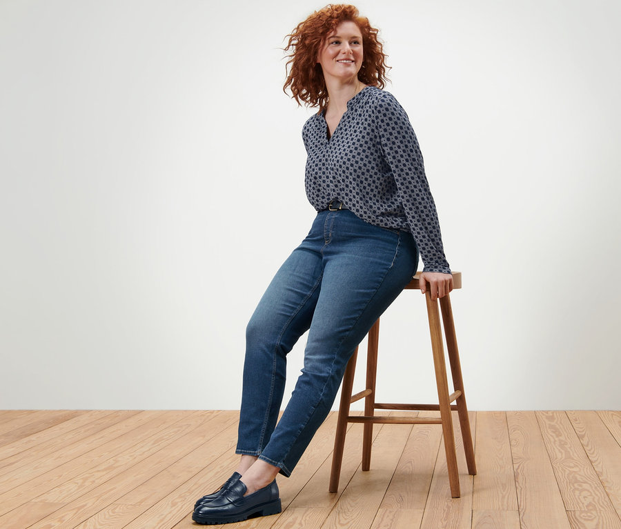 Eine Frau sitzt auf einem Holzhocker und trägt ein bedrucktes Blusenshirt, Treggings »Fit Nelly« und einen Ledergürtel.