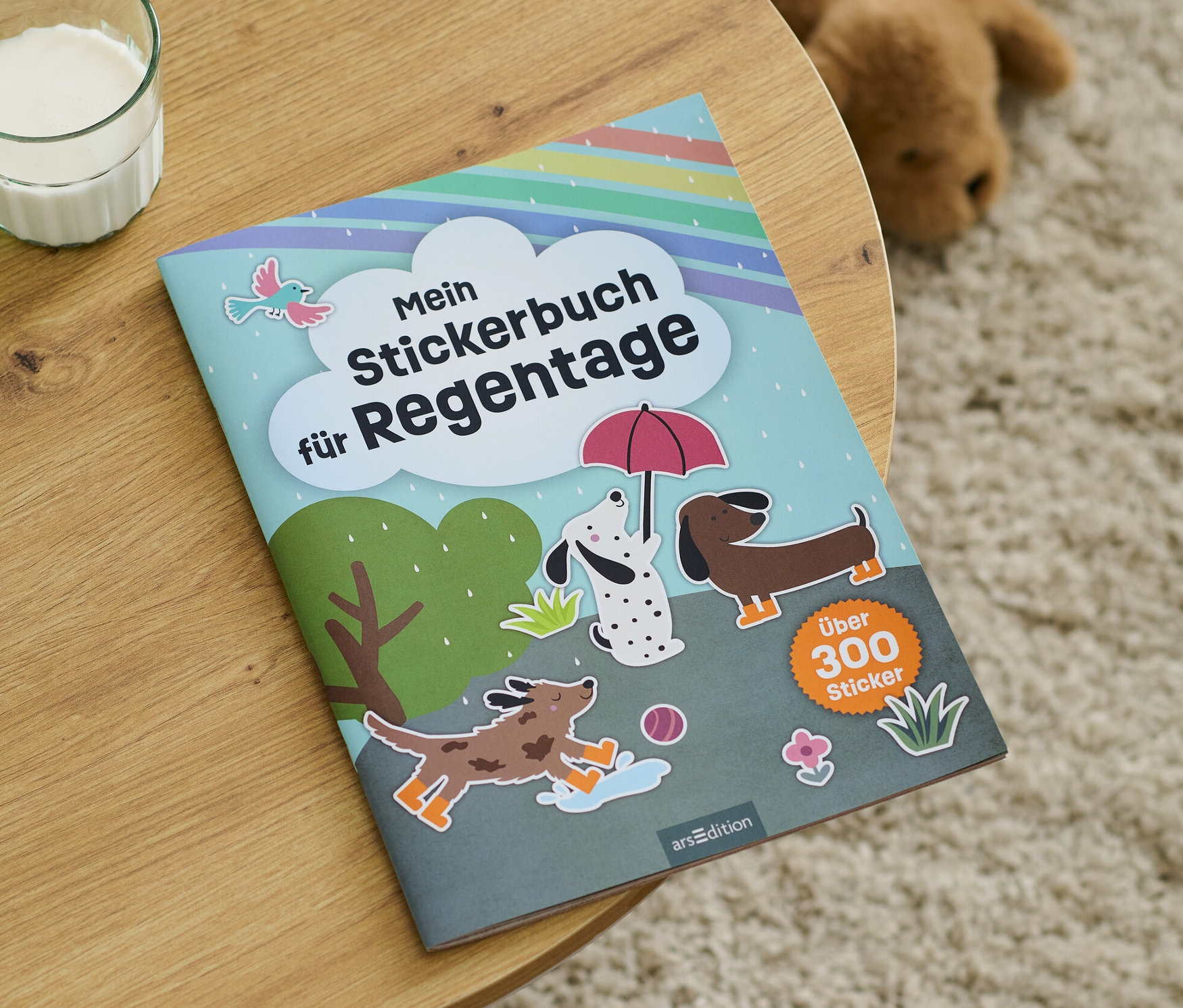 Auf einem Holztisch liegt ein »Mein Stickerbuch für Regentage« neben einem Glas Milch.
