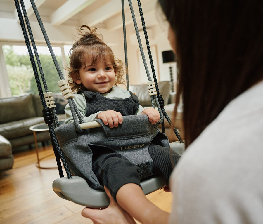 Lächelndes Kind sitzt in grauer HUDORA Schaukelaufsatz Toddler, die von einer erwachsenen Person gehalten wird.