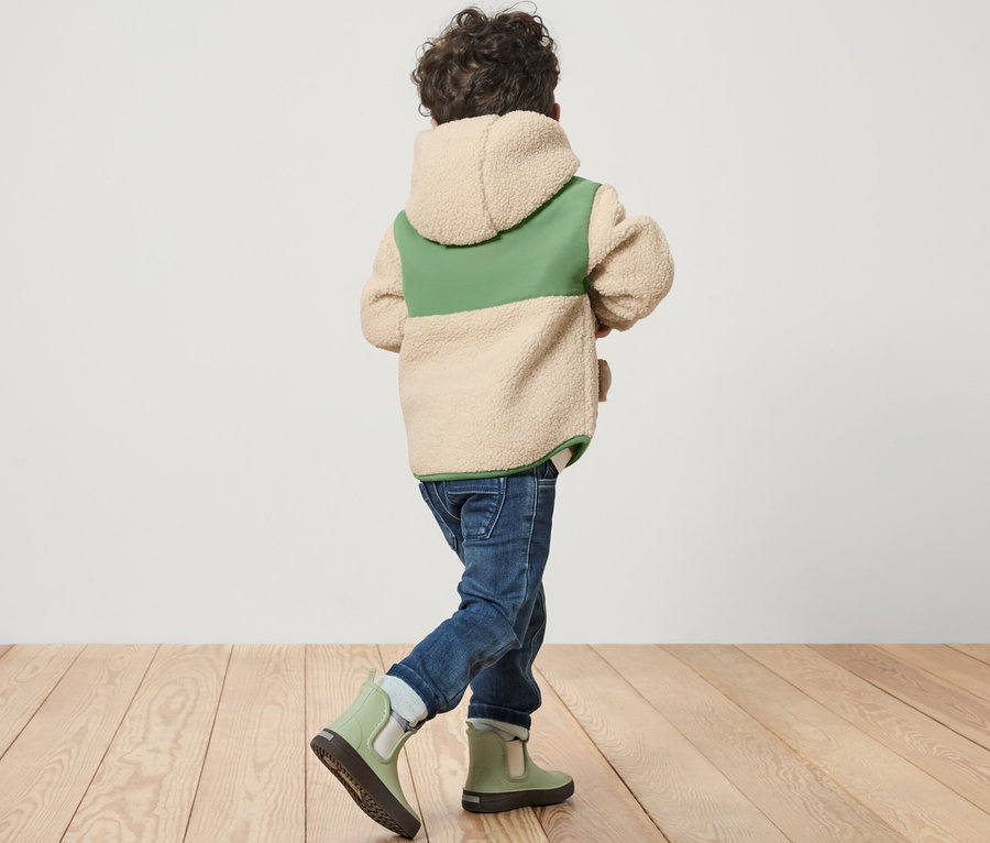 Ein Kind in einer Kinder-Teddy-Jacke geht auf einem Holzboden.