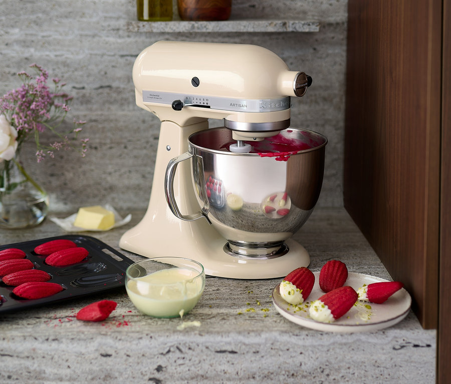 Eine cremefarbene KitchenAid Küchenmaschine »Artisan 5KSM125EAC« rührt rote Glasur in einer Edelstahlschüssel. Auf dem Tisch liegen rote Madeleines, Butter und eine Blumenvase.
