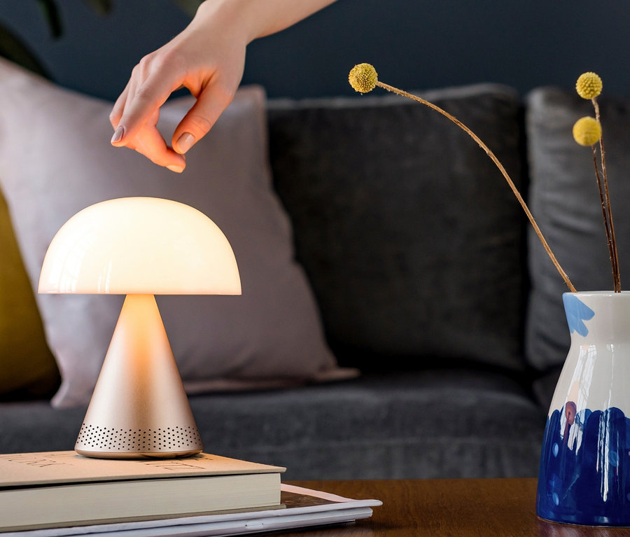 Eine Hand greift nach der goldenen LEXON Akku-Tischleuchte »Mina L Audio«, die auf einem Buch neben einer Blumenvase steht.