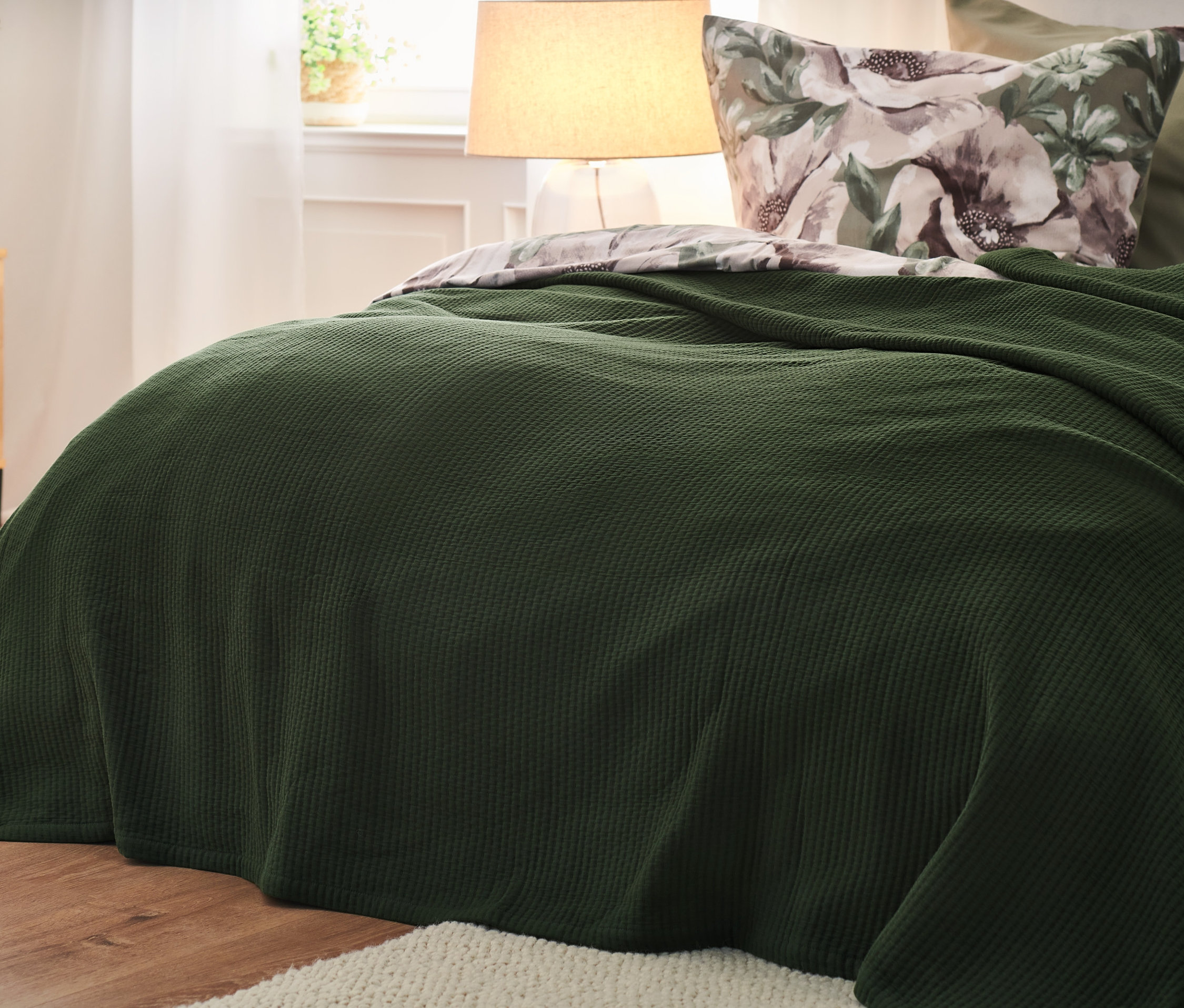 Nahaufnahme einer grünen Bettüberwurf auf einem Bett, im Hintergrund ein Nachttisch und eine Lampe.