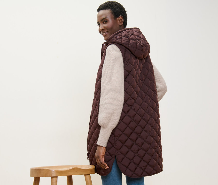 Frau lächelt über die Schulter und trägt eine lange Steppweste mit Kapuze.