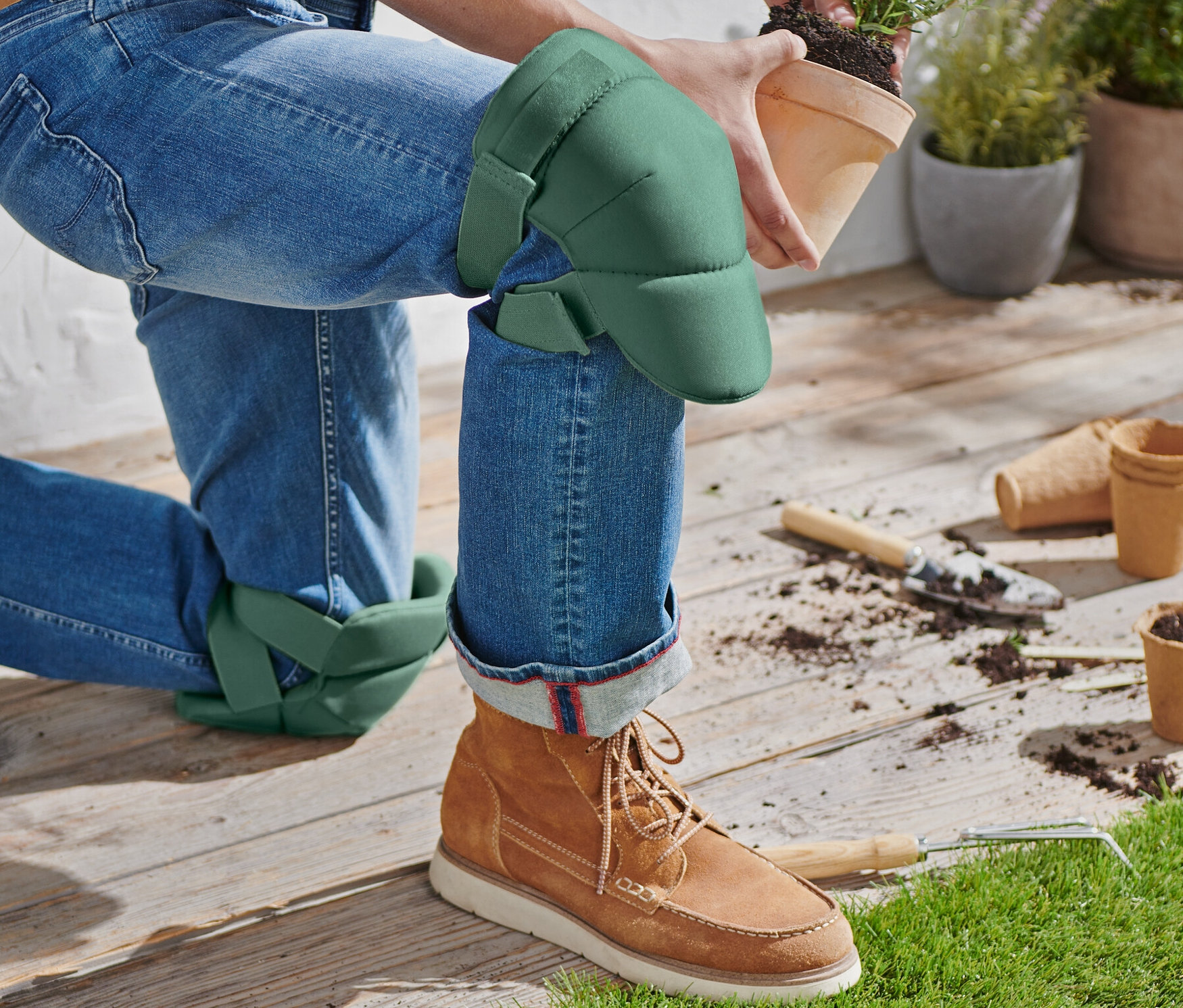 Eine Person kniet mit Garten-Knieschonern und hält einen Topf mit einer Pflanze.