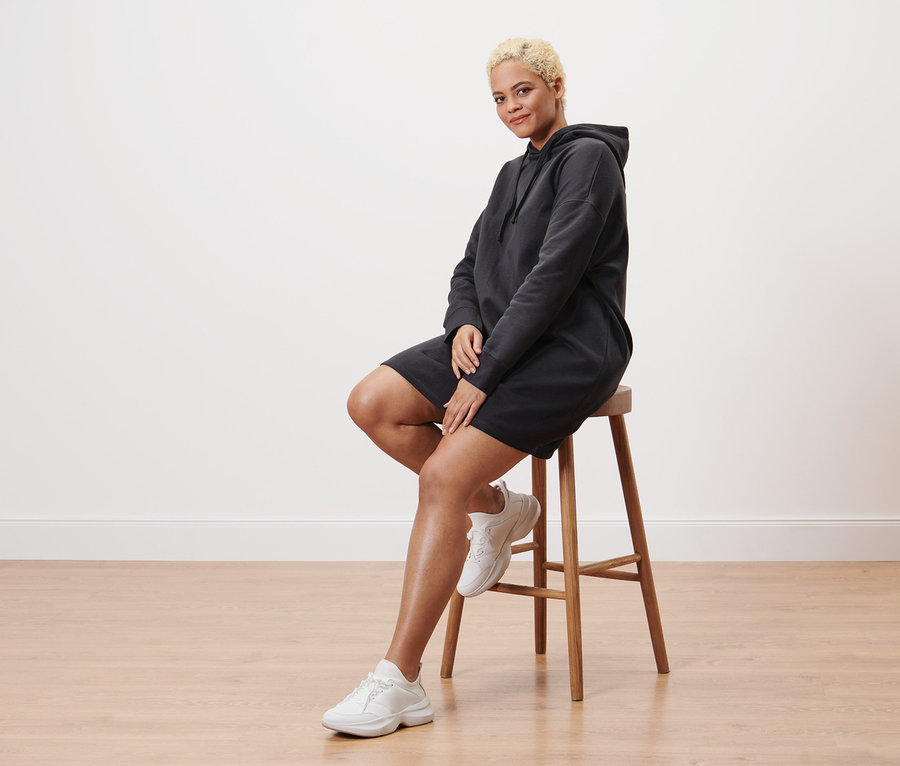 Frau sitzt auf einem Hocker und trägt ein Sweatkleid mit Kapuze.