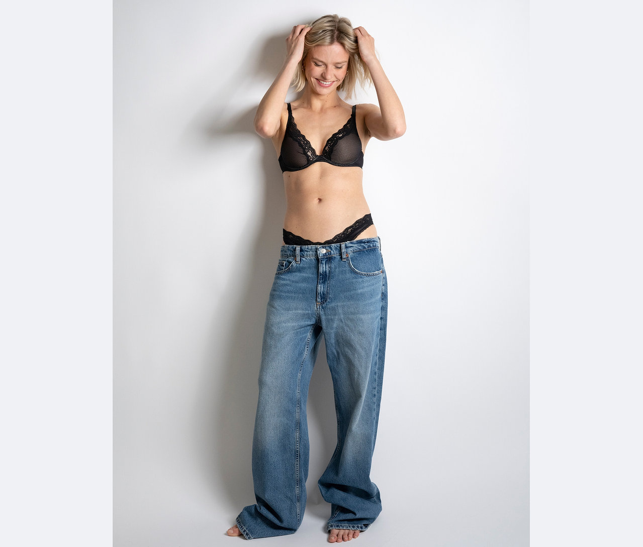 Frau lächelt und hält ihre Hände auf dem Kopf, sie trägt einen schwarzen Spitzen-BH und Tanga und riesige Jeans.