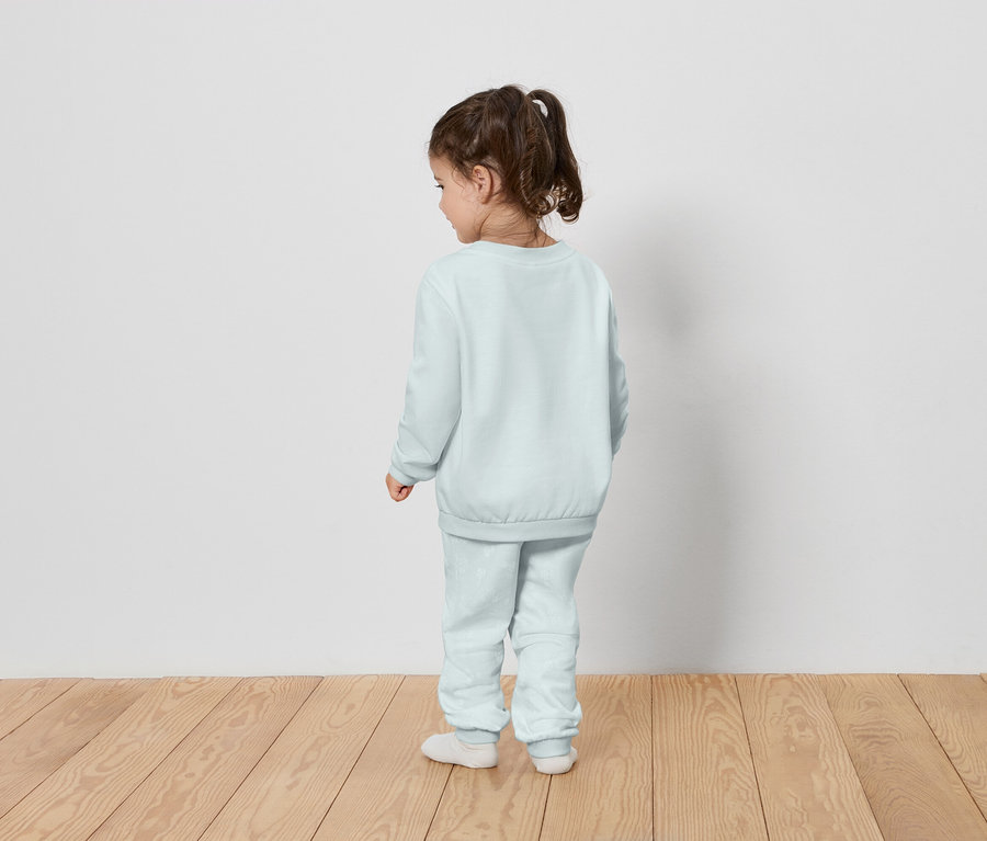 Ein kleines Mädchen steht mit dem Rücken zur Kamera und trägt einen hellblauen Trainingsanzug.