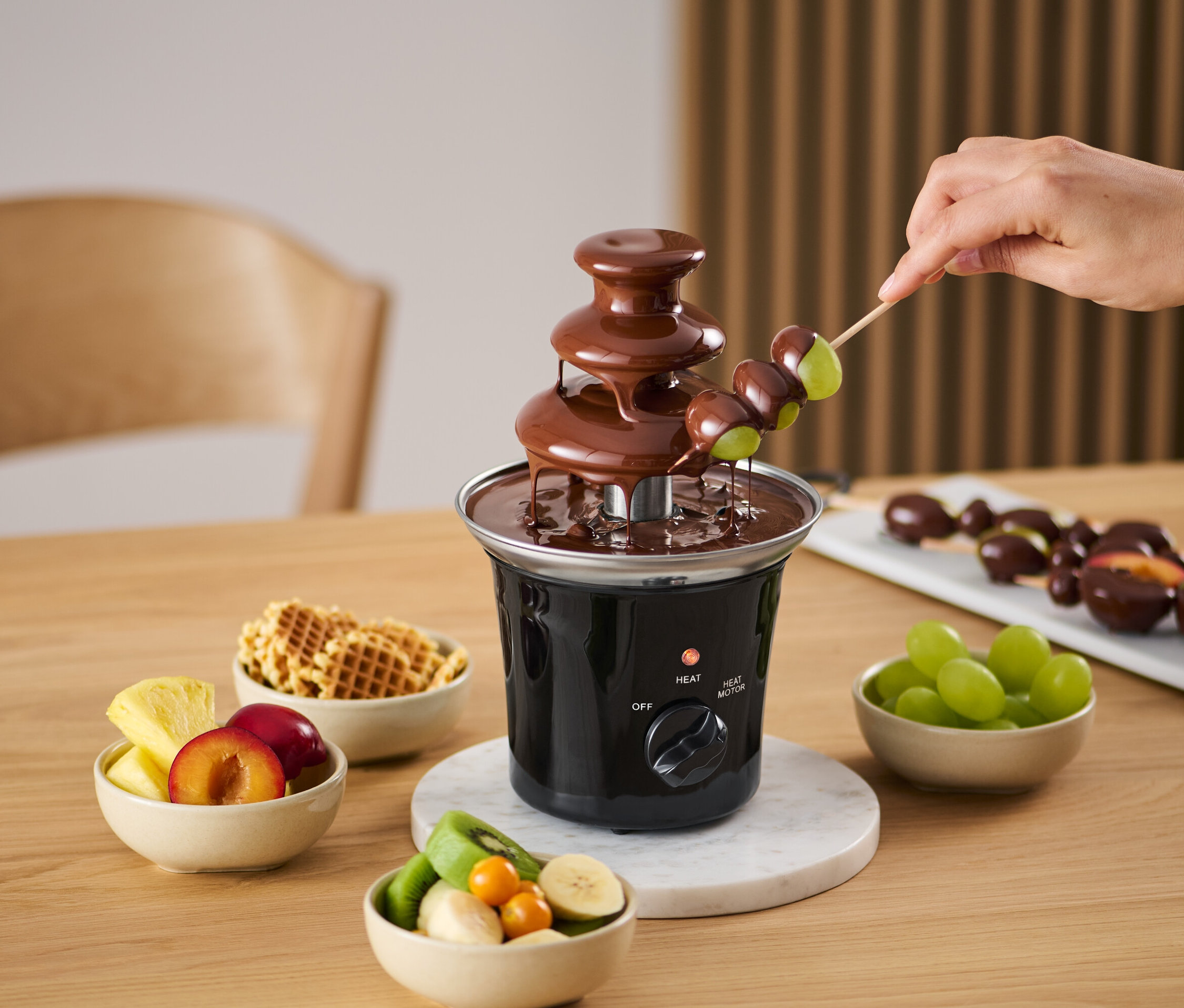 Eine Hand taucht Trauben am Spieß in einen Schokoladenbrunnen. Um den Brunnen herum stehen Schüsseln mit Obst und Waffeln.