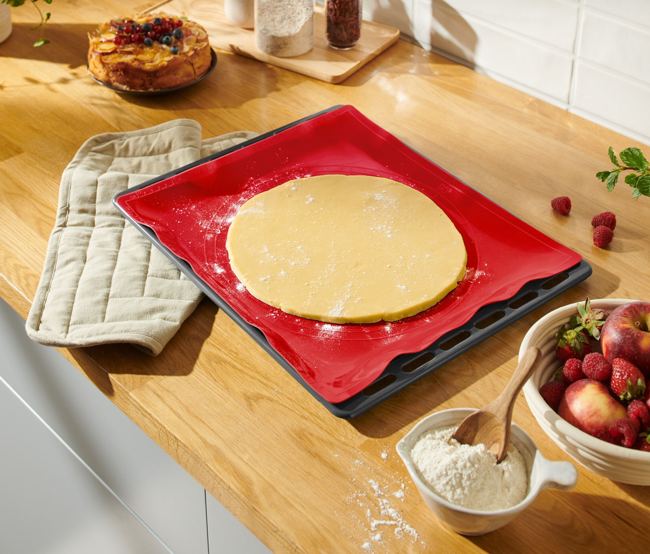 Auf einer Küchentheke liegen eine rote Dauerbackmatte mit Teig, ein Topflappen, eine Schüssel mit Obst und ein Kuchen.
