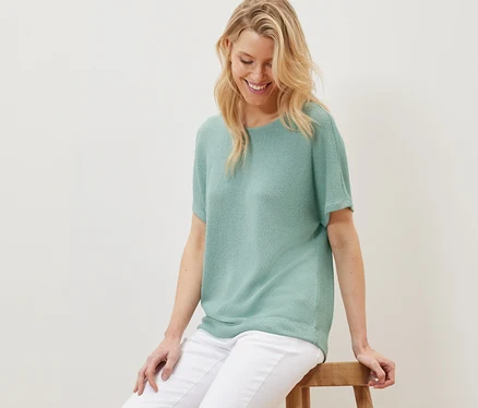 Blonde Frau sitzt auf einem Holzhocker und trägt ein strukturiertes Shirt in Salbei und weiße Hosen.