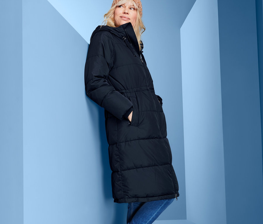 Eine Frau, die sich an eine blaue Wand lehnt, trägt einen Kapuzen-Steppmantel, Jeans, Stiefel und eine Mütze.