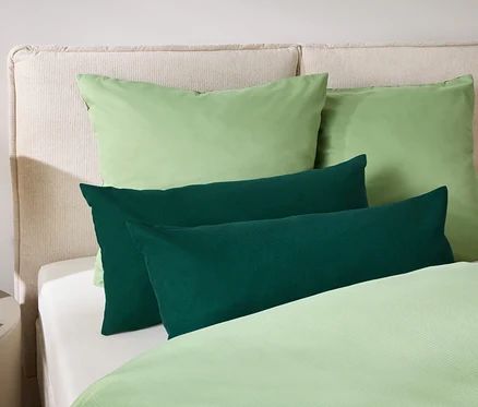 Zwei grüne Kissen und zwei dunkelgrüne längliche Kissen liegen auf einem Bett mit cremefarbenem Kopfteil und hellgrüner Bettwäsche.