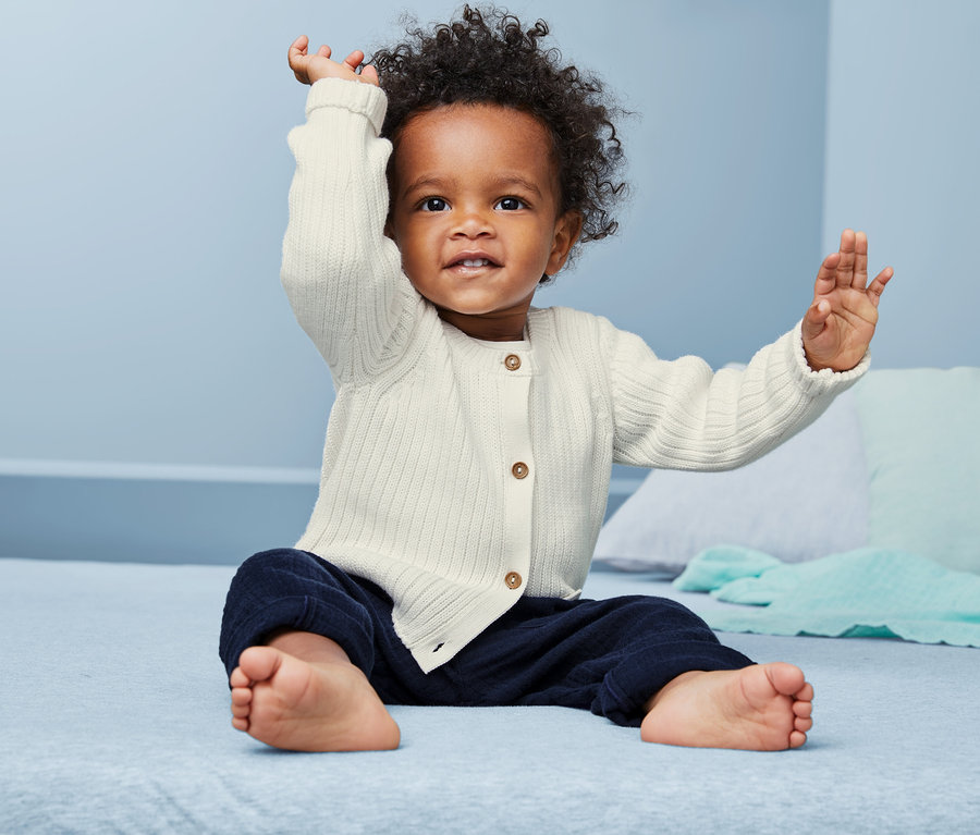 Sitzendes Baby mit erhobenen Armen, trägt eine Baby Baumwoll-Strickjacke.