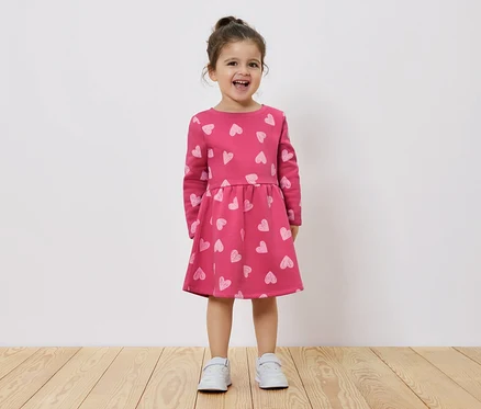Ein lächelndes kleines Mädchen steht auf einem Holzboden und trägt ein rosa Kleid mit langen Ärmeln und Herzchenmuster.