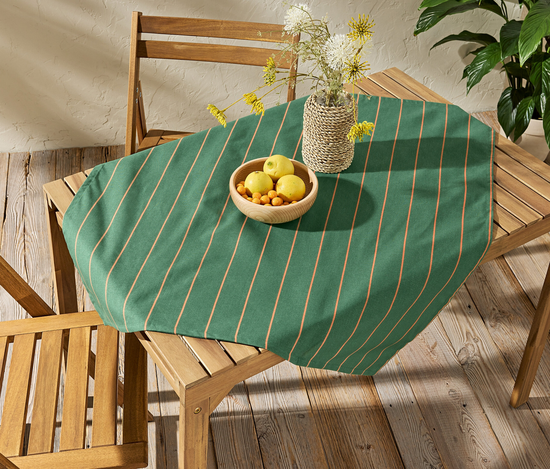 Auf einem Holztisch liegt eine abwischbare Tischdecke, ca. 90 x 90 cm, gestreift. Auf der Tischdecke stehen eine Schale mit Obst und eine Vase mit Blumen.