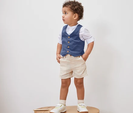 Kleiner Junge steht auf einem Holzhocker und trägt eine Kinder-Weste, ein Hemd, Shorts und Schuhe.