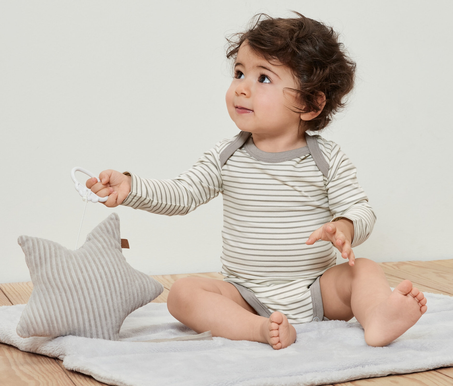 Lockiges Baby sitzt auf einer Decke und spielt mit einem Stern. Es trägt 5 Baby-Bodys mit Mitwachsfunktion.