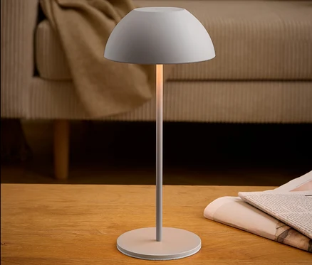Eine graue Lampe steht neben einer Zeitung auf einem Holztisch. Im Hintergrund befindet sich ein beiges Sofa.