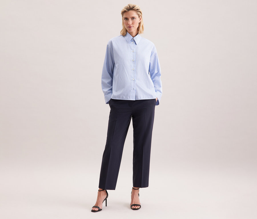 Frau trägt eine Seidensticker Oversize-Hemdbluse mit blauen Streifen, dunkle Hose und schwarze Sandalen mit Absatz.