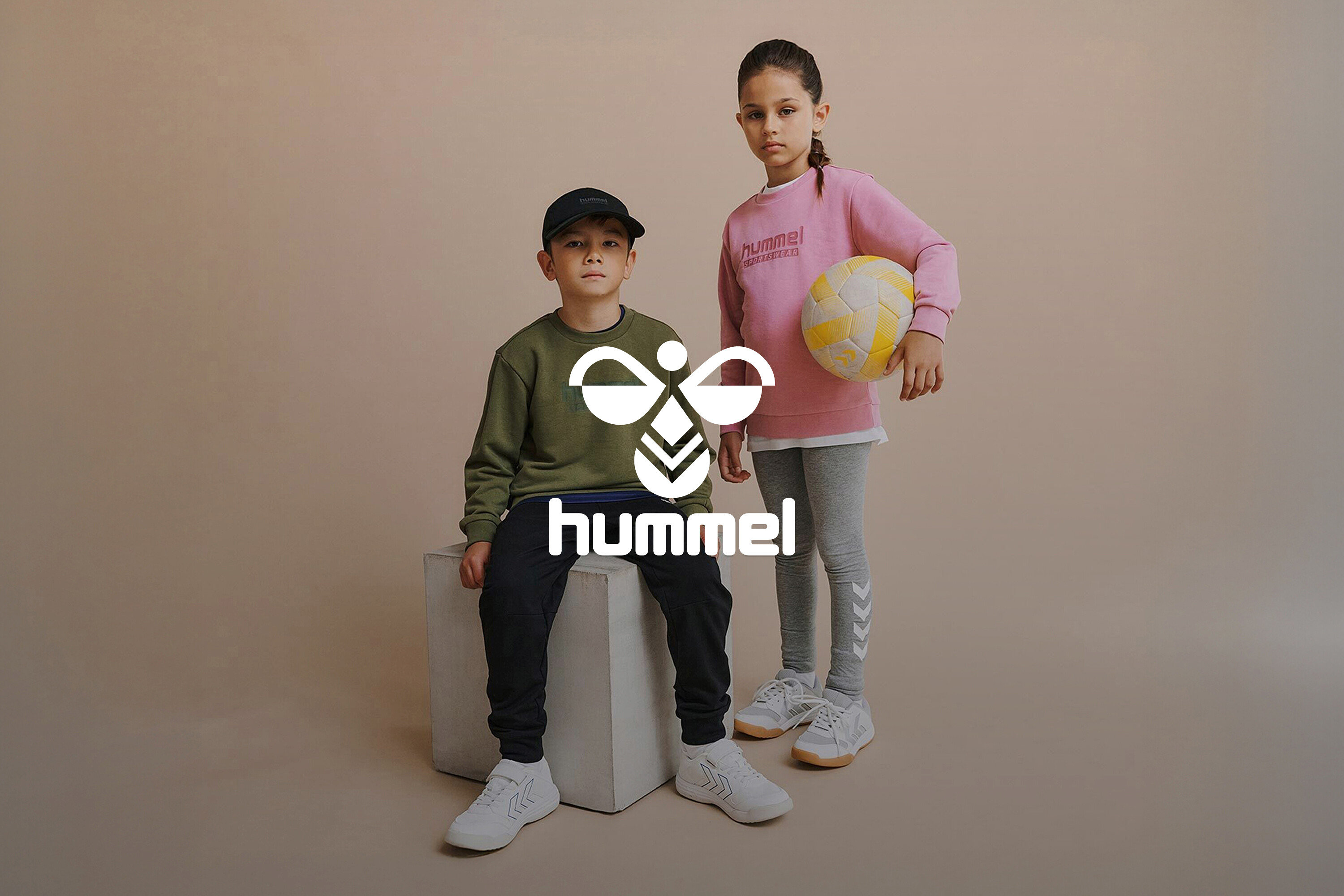 Ein Junge in olivgrünem Sweatshirt sitzt auf einem weißen Würfel, ein Mädchen in einem rosa Sweatshirt hält einen gelb-weißen Ball.