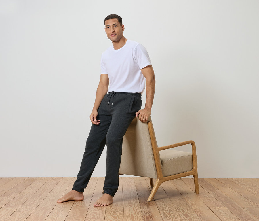 Mann lehnt an einem Stuhl, trägt ein weißes T-Shirt und graue Trainingshose.