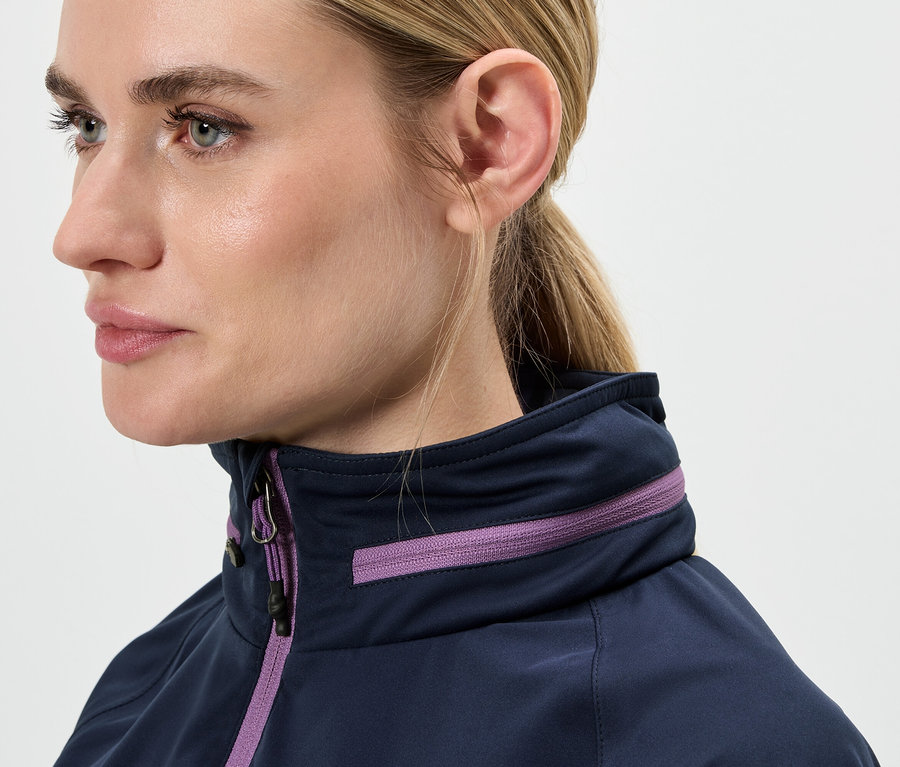 Detailaufnahme des Gesichts einer Frau mit blonden Haaren, die zu einem Pferdeschwanz zusammengebunden sind, und einer blauen Jacke mit lila Details.