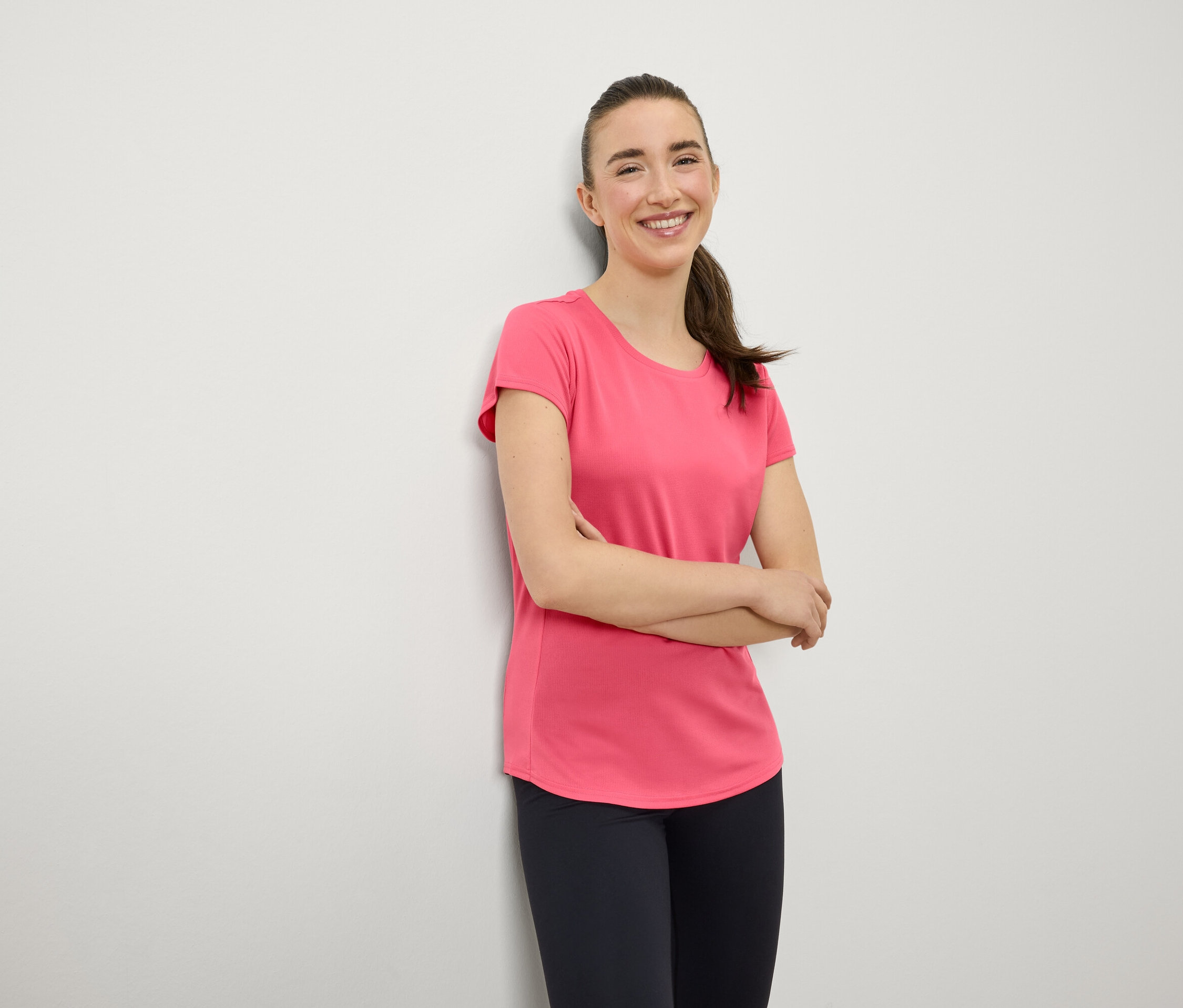 Eine lächelnde Frau mit braunen Haaren im Pferdeschwanz steht mit verschränkten Armen vor der Brust. Sie trägt ein rosa T-Shirt und marineblaue Leggings.